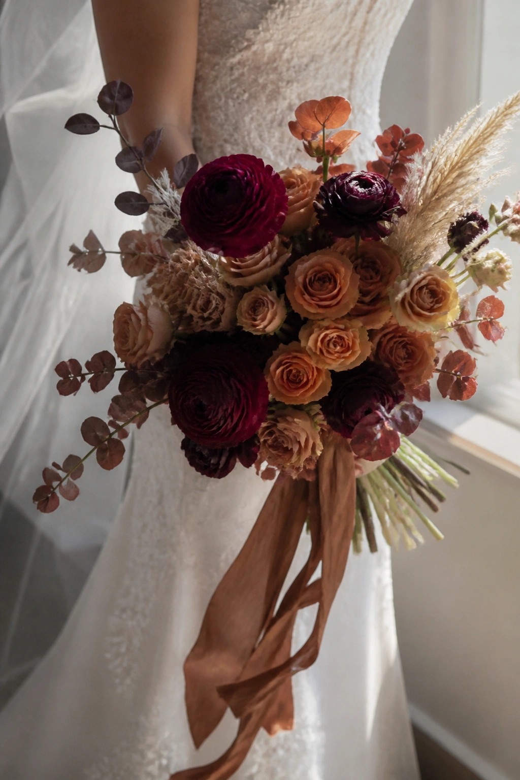 Close up of a bride in a white lace dress holding a fall bouquet of burgundy ranunculus, copper roses, dried grasses, and rust eucalyptus tied with brown ribbons.