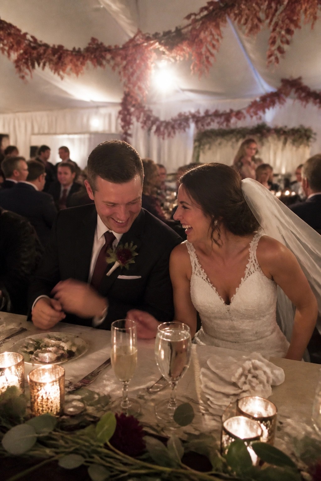 Bride and groom laughing at reception table under hanging burgundy leaves with candlelight and fall decor.