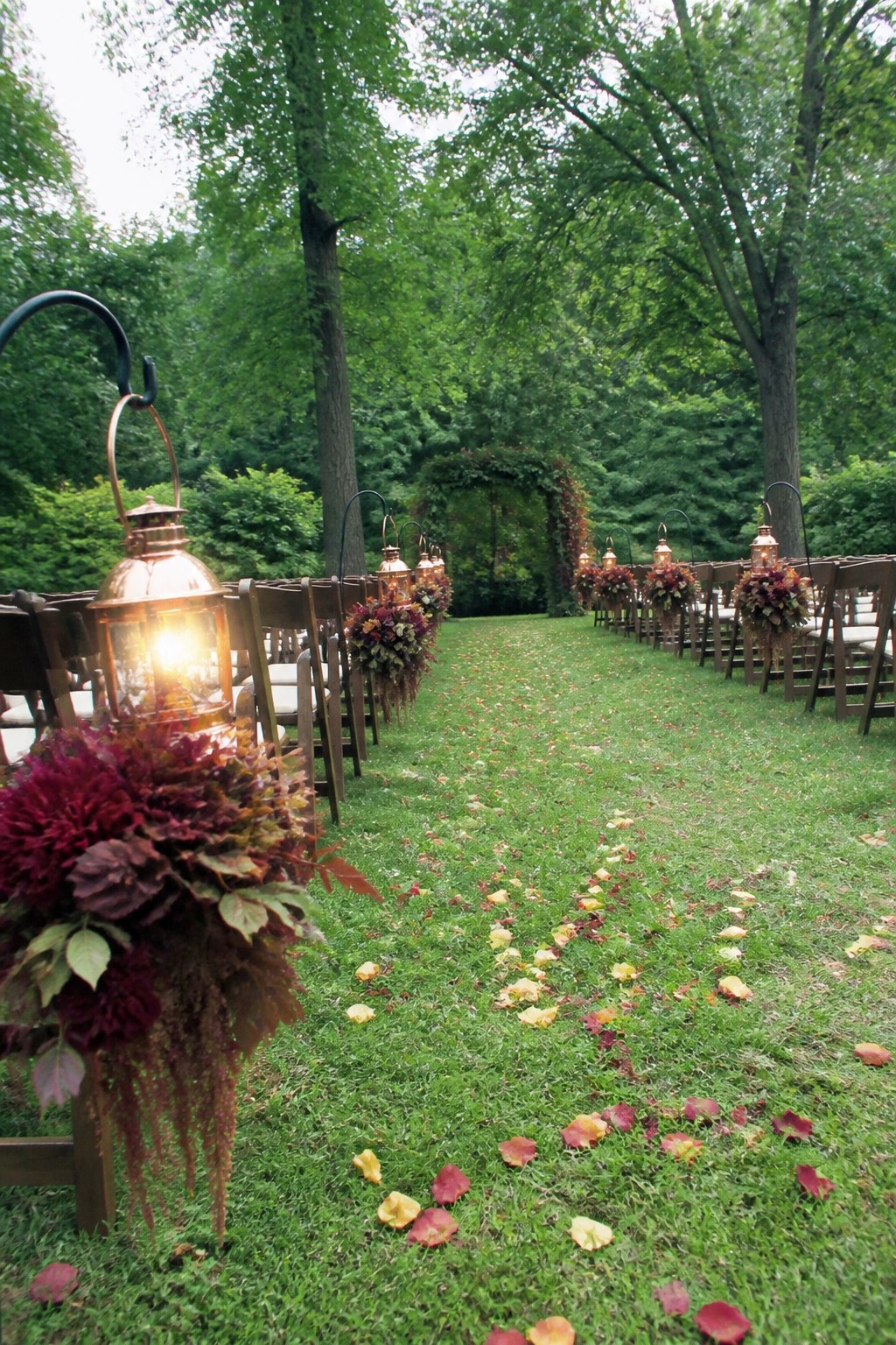 Outdoor fall wedding aisle lined with wooden chairs adorned with burgundy floral arrangements and glowing copper lanterns on a grass path scattered with autumn leaves in a lush forest.