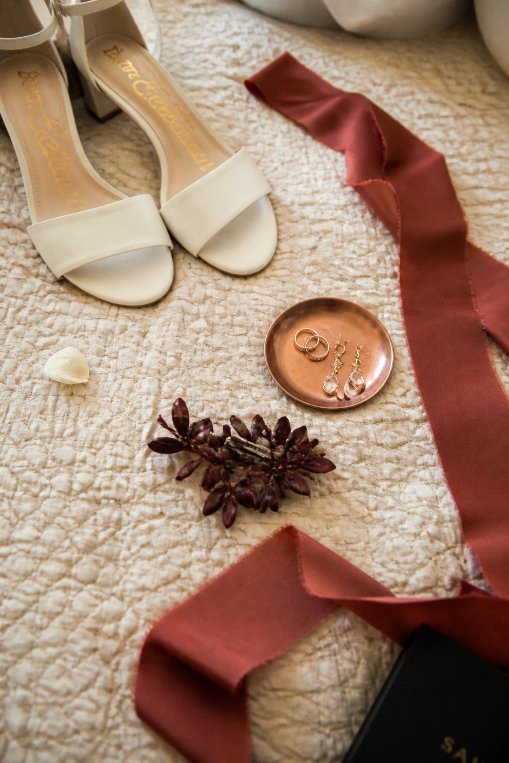 White Sam Edelman heels beside a copper tray of rings and earrings with burgundy ribbons and foliage on a quilted cream background