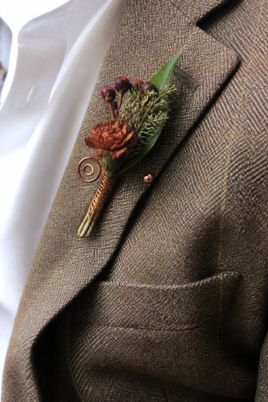 Close up of a rust flower boutonniere with burgundy berries and copper wire on a brown tweed jacket lapel at a fall wedding.