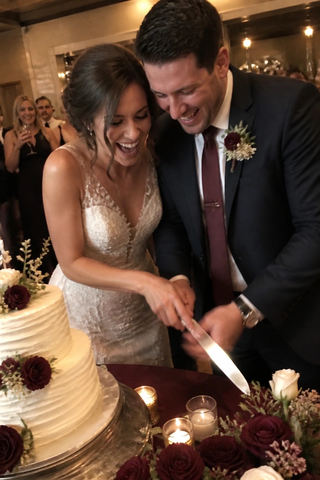 Bride in lace gown and groom in navy suit cut white wedding cake decorated with burgundy roses while smiling at each other during reception.