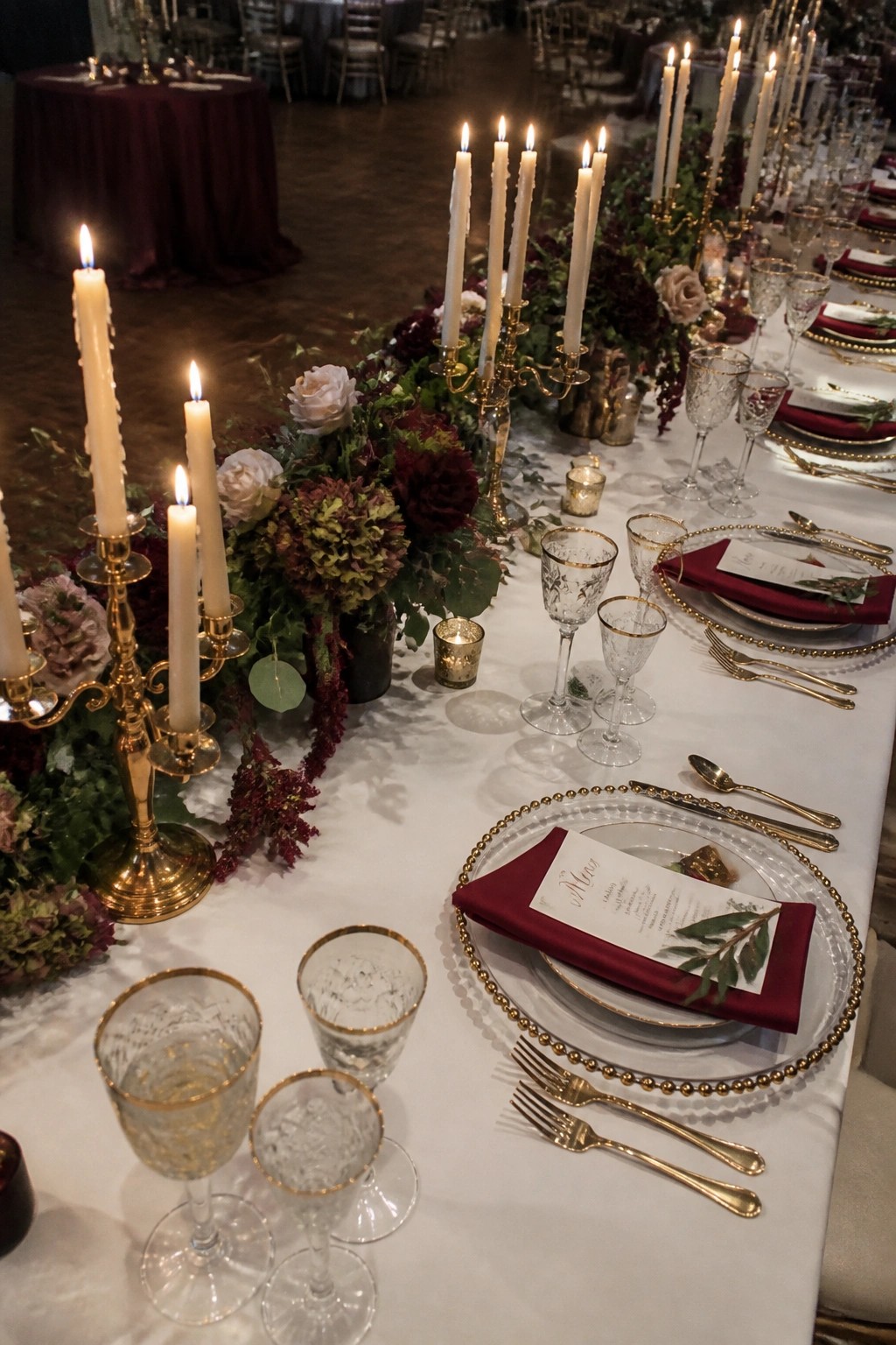 Long wedding table with gold candelabras, white candles, burgundy and white florals, crystal glasses, and burgundy napkins on white linens.