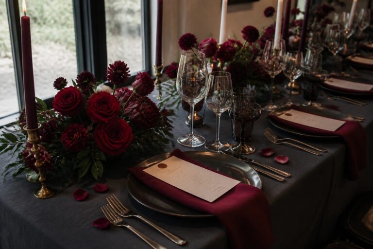 Formal wedding table with white plates, burgundy napkins, gold cutlery, dark glassware, tall burgundy candles, and lush burgundy floral arrangements on a dark gray tablecloth.