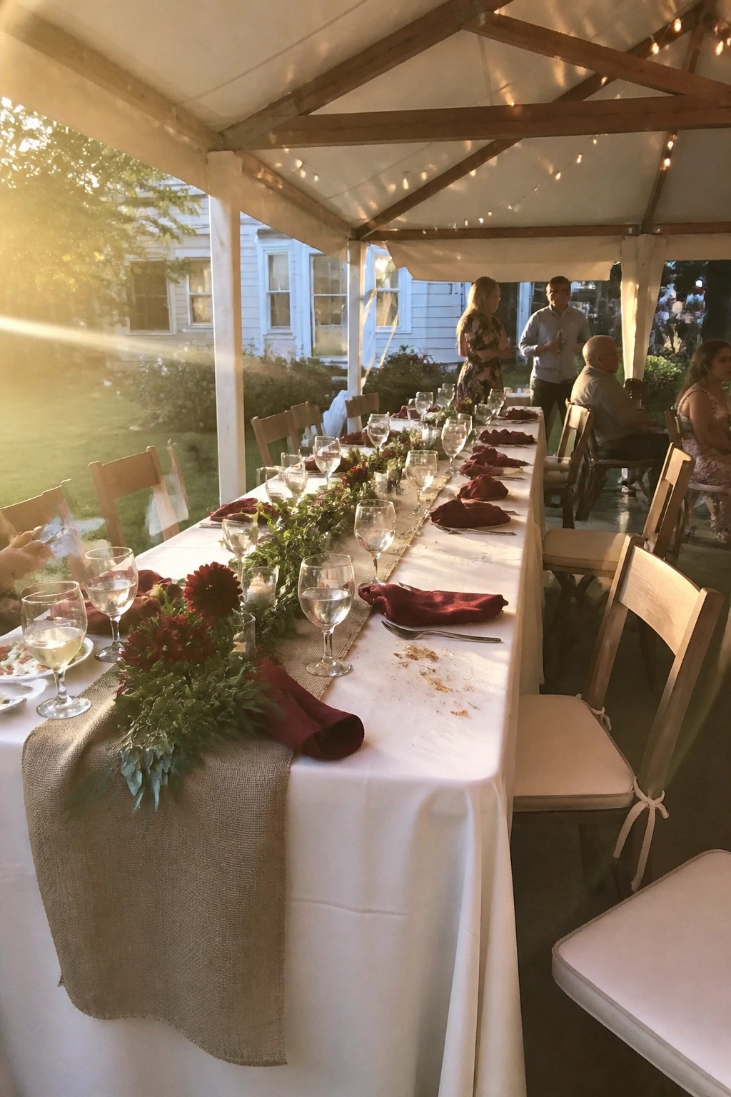 Long wedding table under tent with white cloth, burlap runner, burgundy napkins, red dahlias, wine glasses, and string lights at sunset.