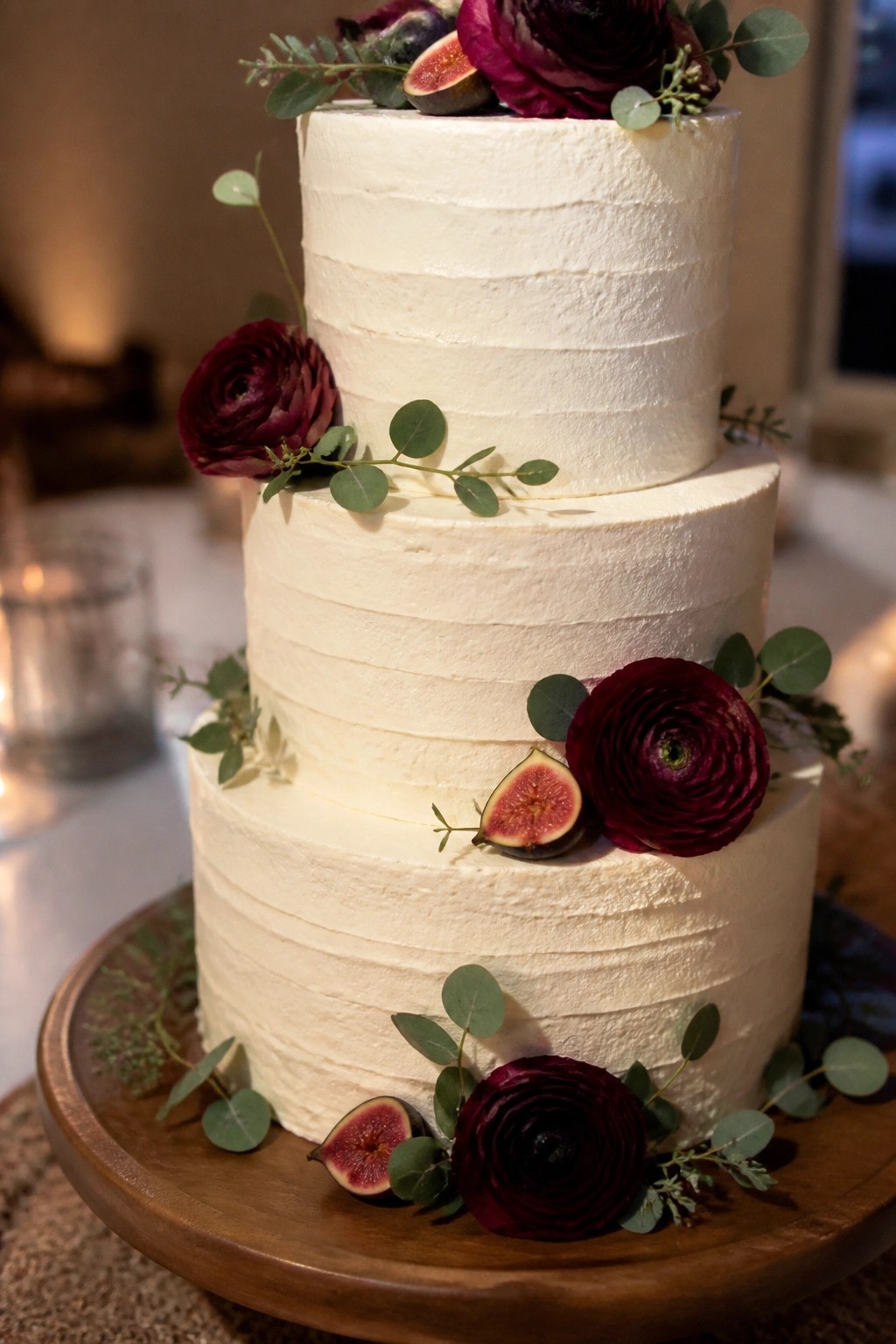 Three tier white wedding cake with burgundy ranunculus flowers figs and eucalyptus on a wooden stand in soft lighting