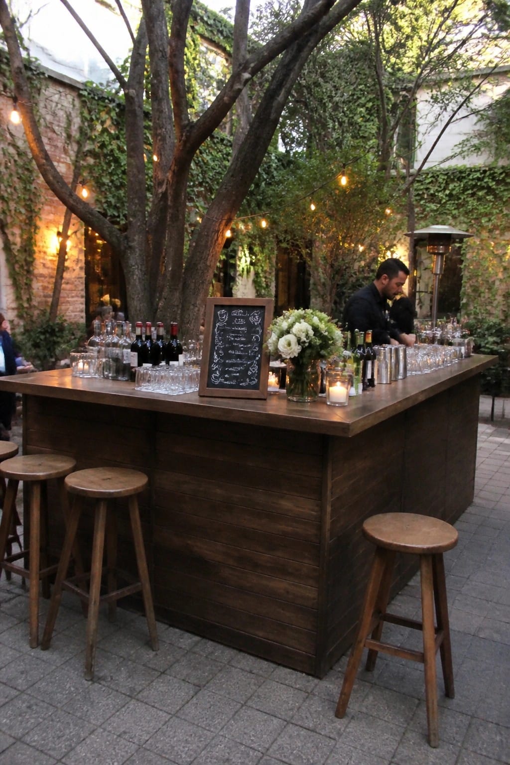 Rustic wooden bar in a garden courtyard with string lights wooden stools chalkboard menu and floral arrangements at a wedding reception