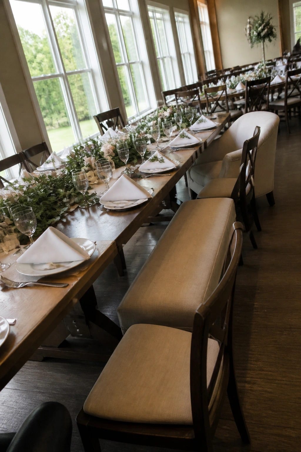 Long wooden wedding table set with white linens and floral garland beside large windows with mixed upholstered and wooden chairs in a bright hall.