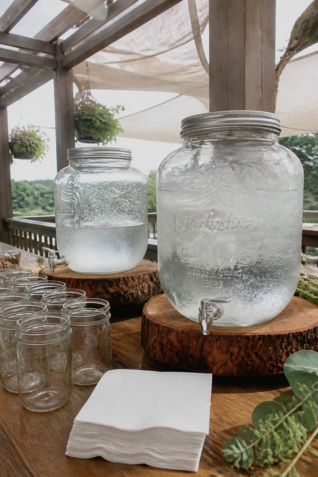 Two large glass drink dispensers with spigots on wooden stands filled with ice water beside rows of empty mason jars and white napkins on an outdoor wooden table at a wedding reception