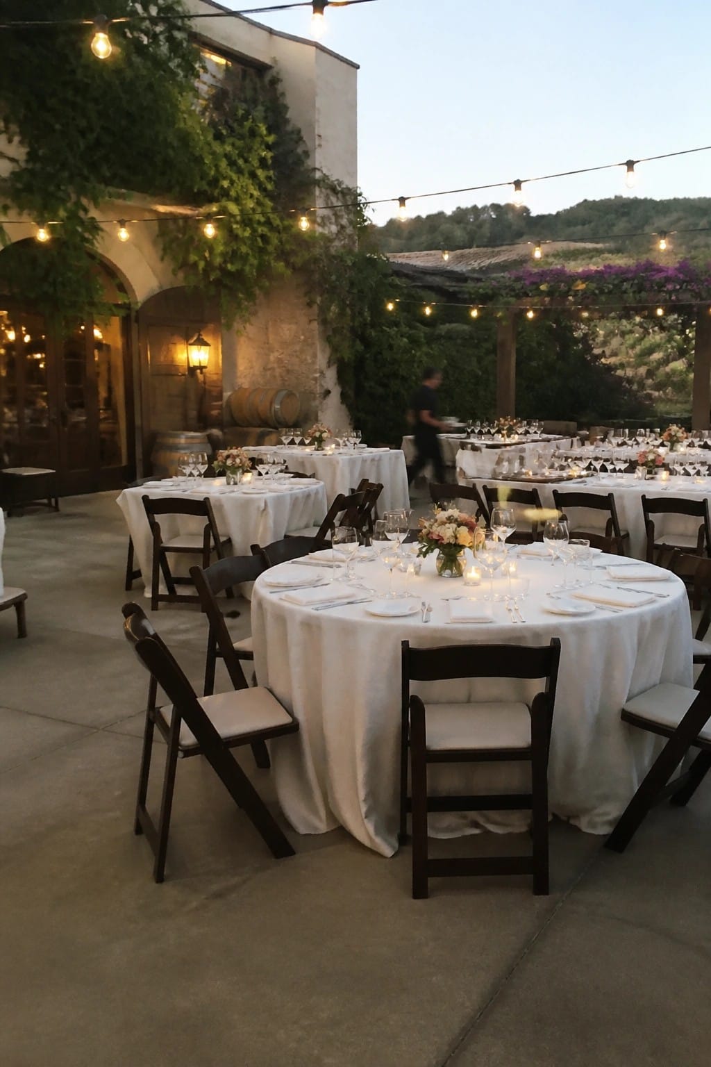 Outdoor wedding reception at dusk with round tables in white linens, dark wooden chairs, string lights, floral centerpieces, and a stone building with greenery.