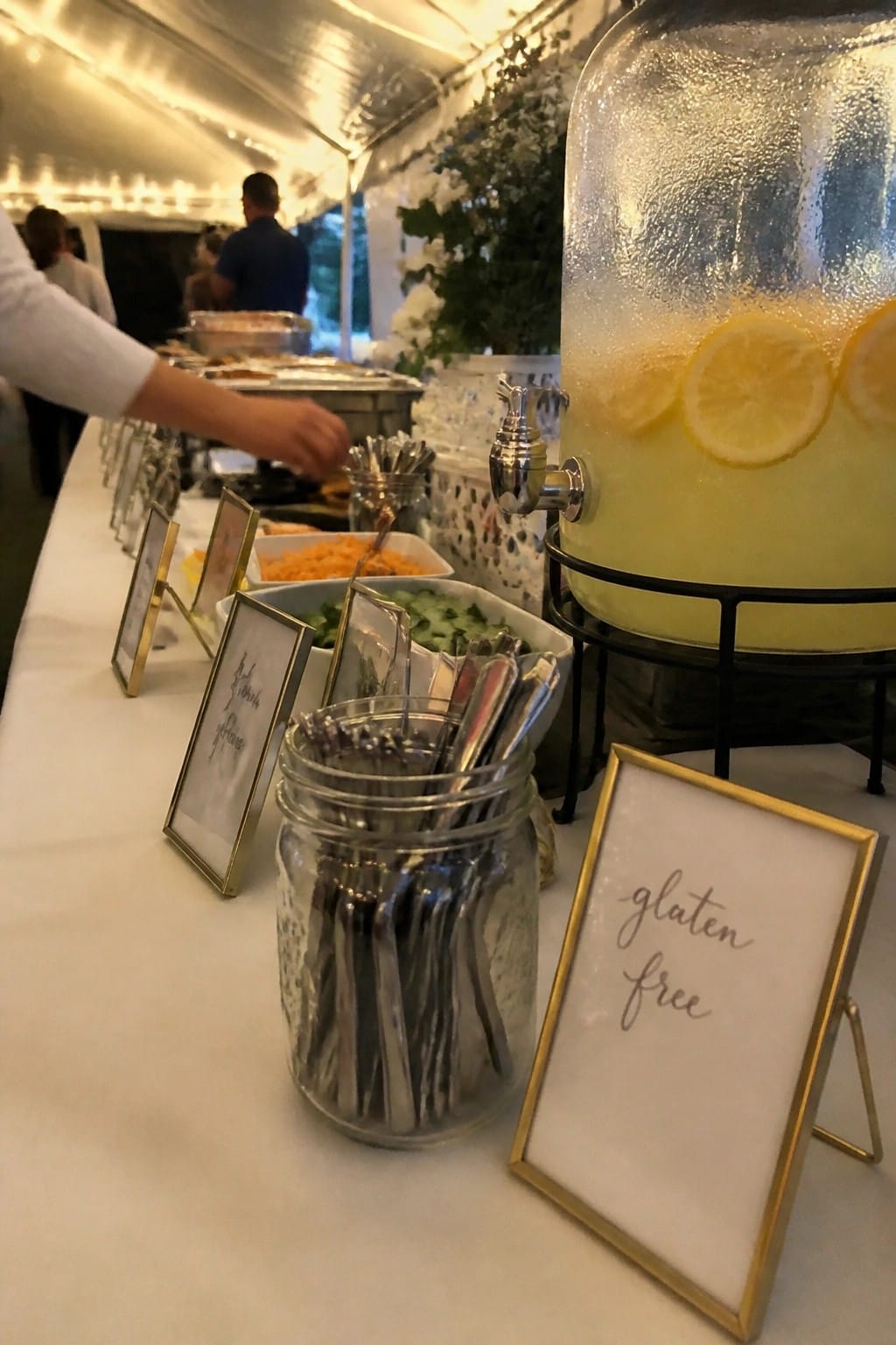 A wedding buffet table displays a lemonade dispenser with lemon slices and a framed gluten free sign beside jars of utensils and labeled food dishes under tent lighting.