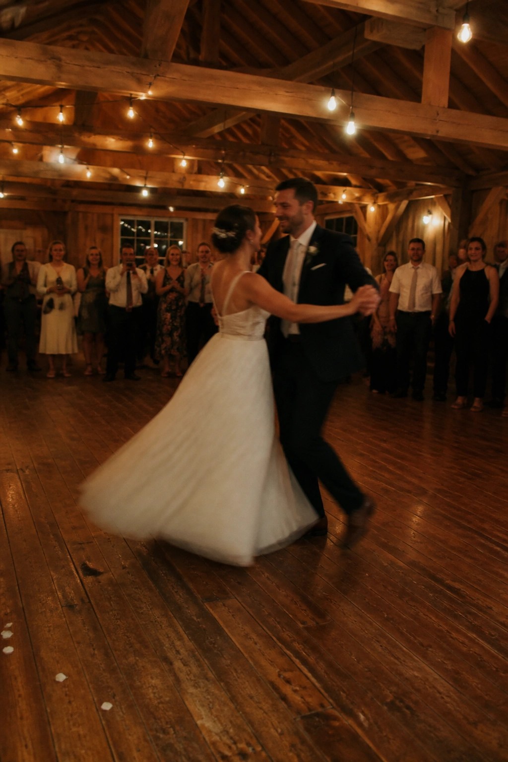 Bride in flowing white gown and groom in dark suit dance together in a rustic wooden barn under glowing string lights with guests watching in the background.