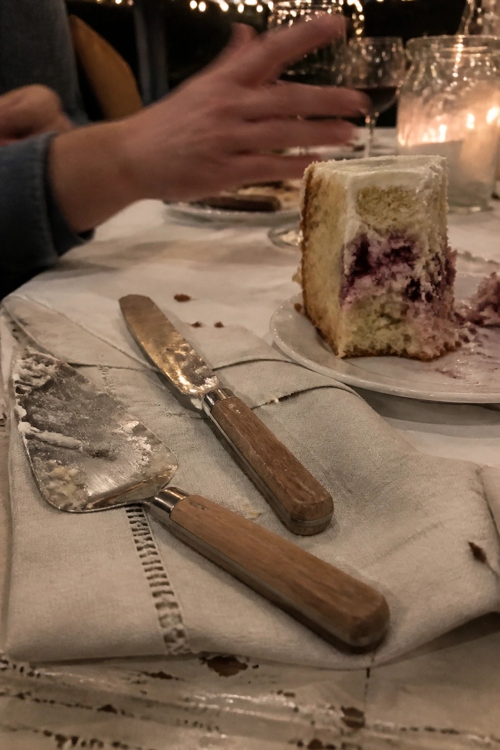 A close up of a berry filled cake slice on a plate with wooden cake servers on a napkin and a lit candle glowing nearby at a rustic wedding reception.