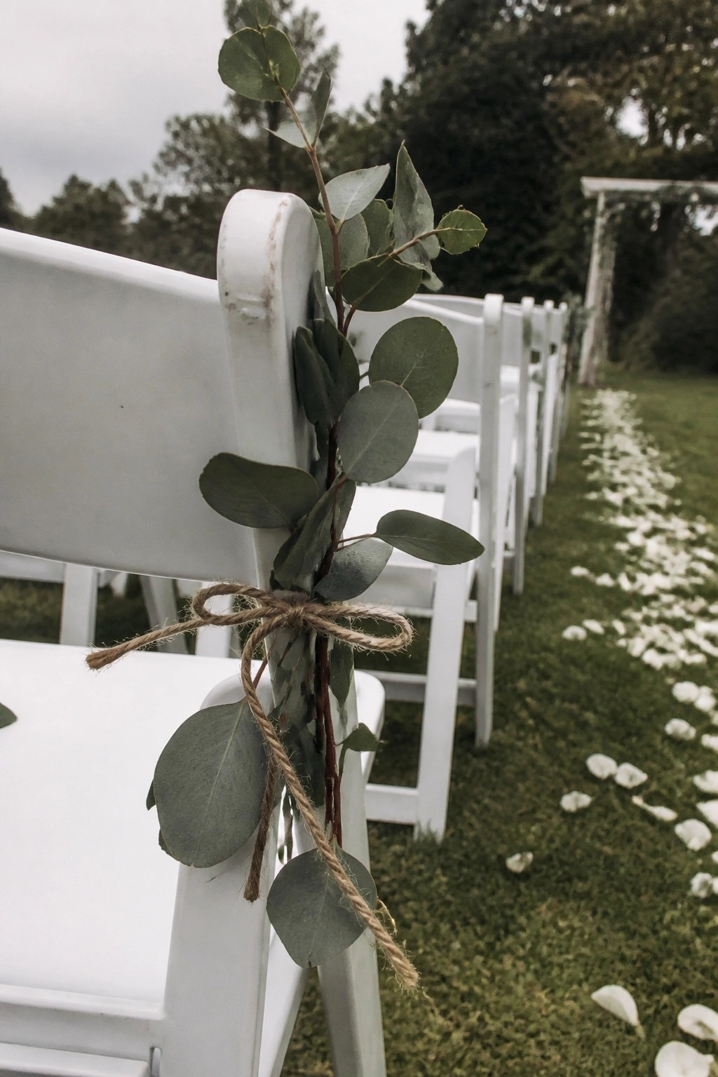 Close up of a white folding chair with eucalyptus leaves tied by twine on the back next to a petal lined aisle in an outdoor wedding setting