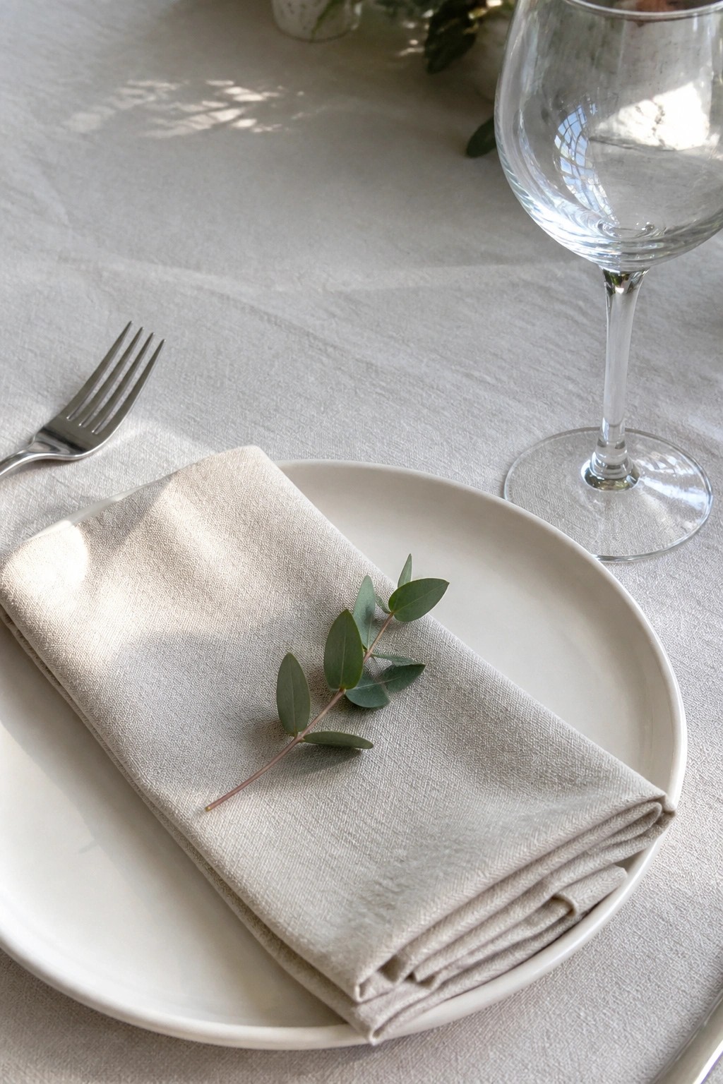 Minimalist wedding place setting with white plate beige linen napkin eucalyptus sprig fork and wine glass on white tablecloth in soft natural light