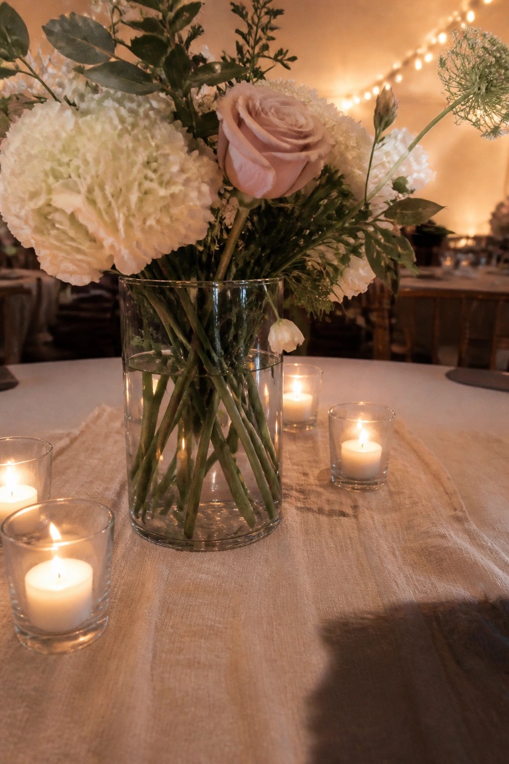 A glass vase with white hydrangeas and a pale pink rose surrounded by glowing tea lights on a linen runner at a wedding reception table.