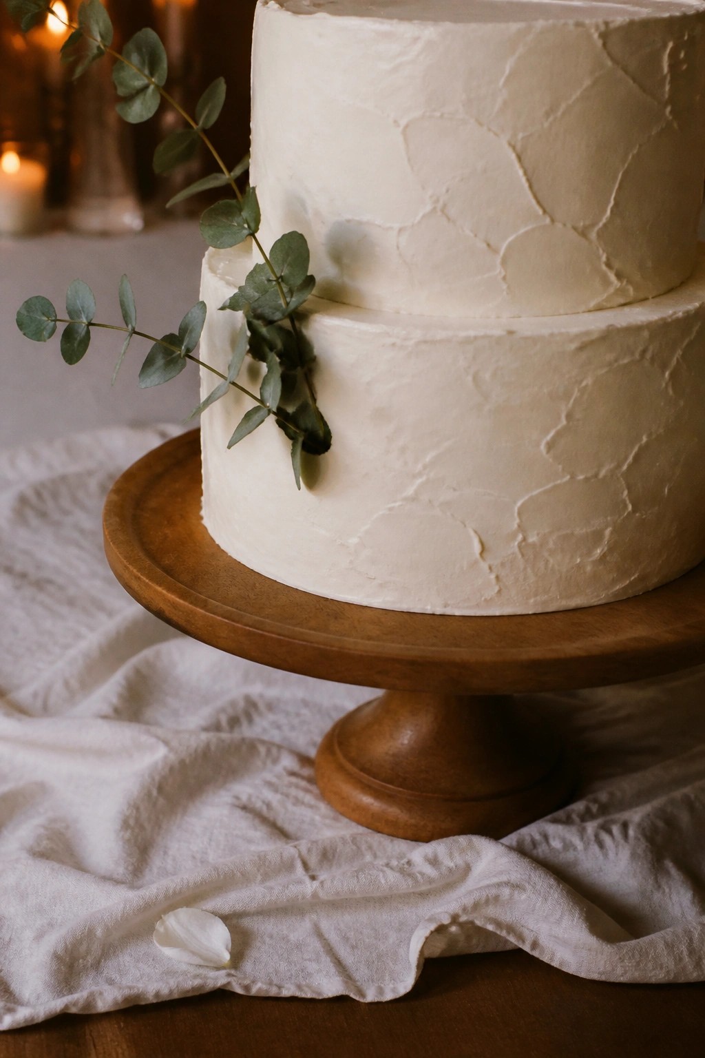 Two tier white textured wedding cake on wooden stand with eucalyptus greenery against warm candlelit background on white linen cloth.