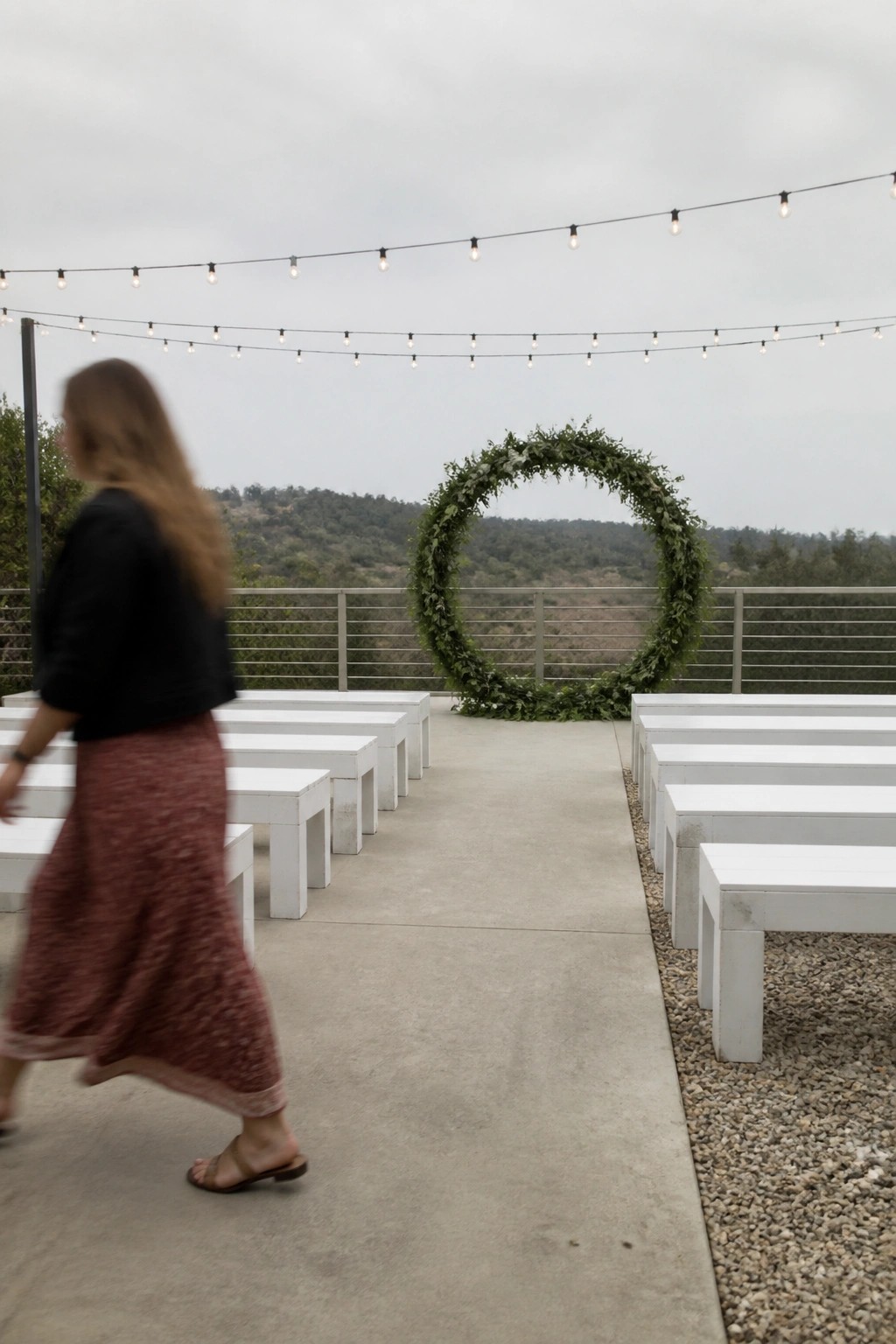 Outdoor wedding terrace with white benches in rows, a large circular greenery arch at the end, string lights above, and a scenic hill view under gray skies.