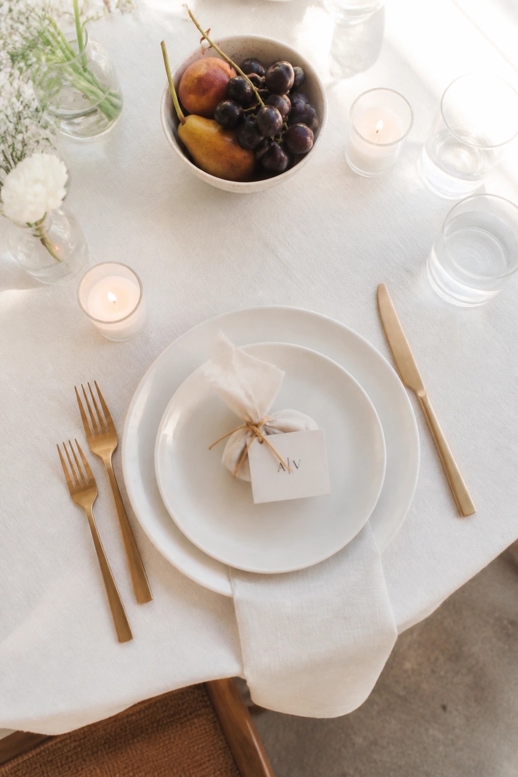 Top down view of a minimalist wedding place setting with white plates gold cutlery fruit bowl and candles on a white tablecloth