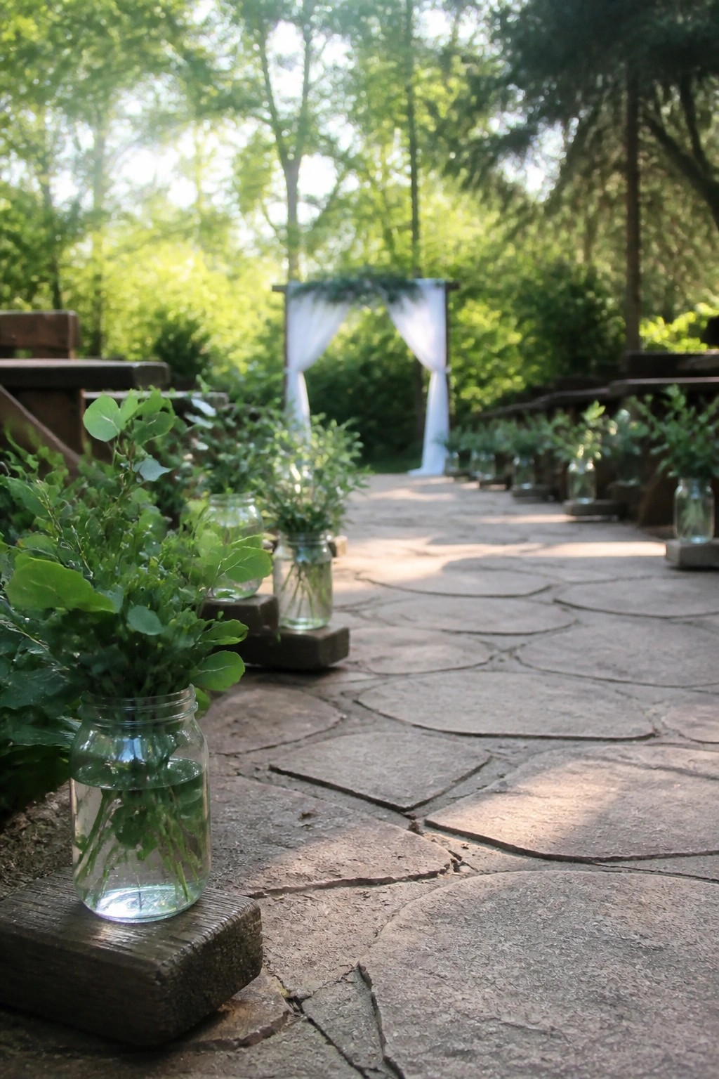 Stone pathway lined with mason jars of green foliage leading to a white draped wedding arch in a forest setting