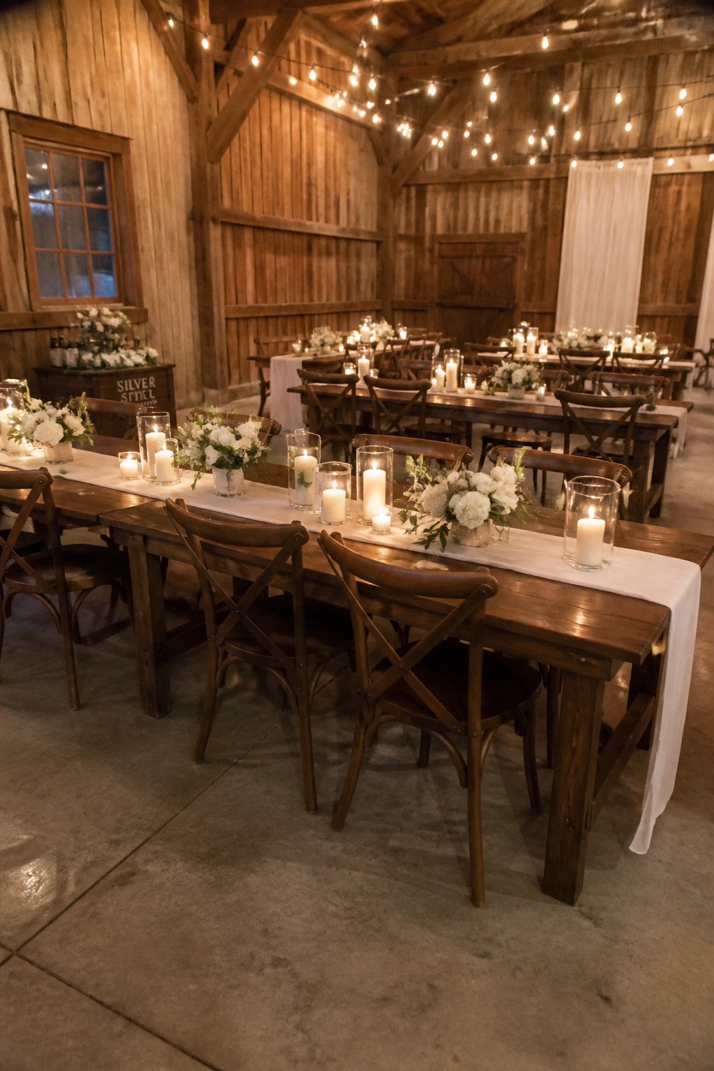 Long wooden tables in a rustic barn wedding venue feature white runners, glowing candles in glass holders, and white floral arrangements under string lights.