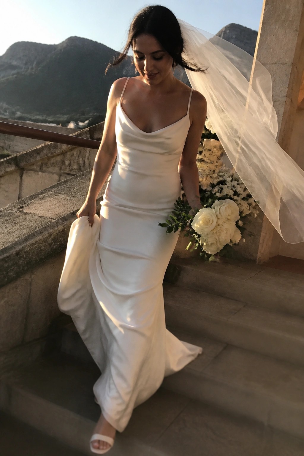Bride in minimalist white silk gown on stone steps holding white rose bouquet with flowing veil and mountain backdrop at sunset