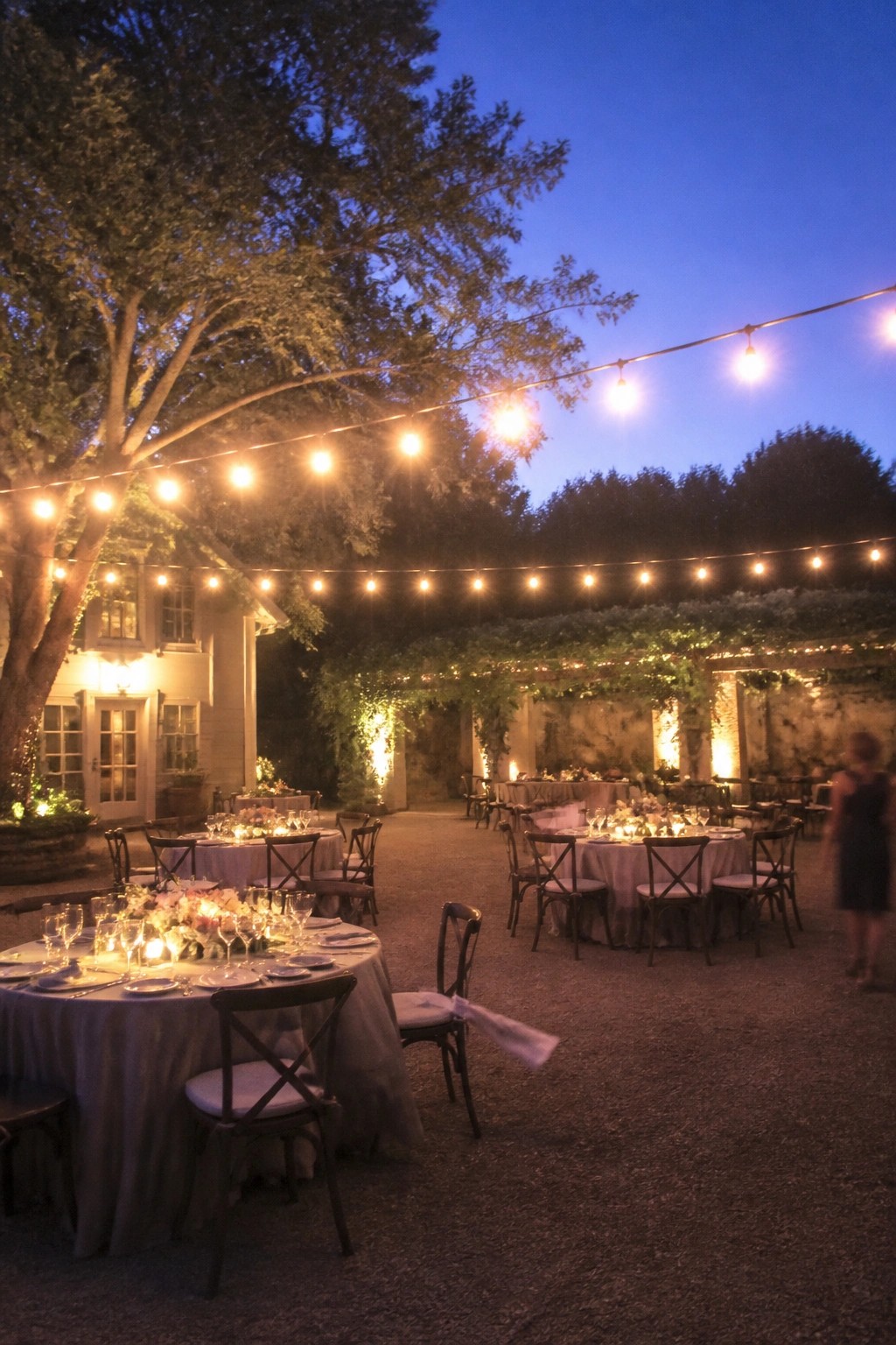 Evening outdoor wedding reception with round tables under glowing string lights a large tree and stone wall at dusk.