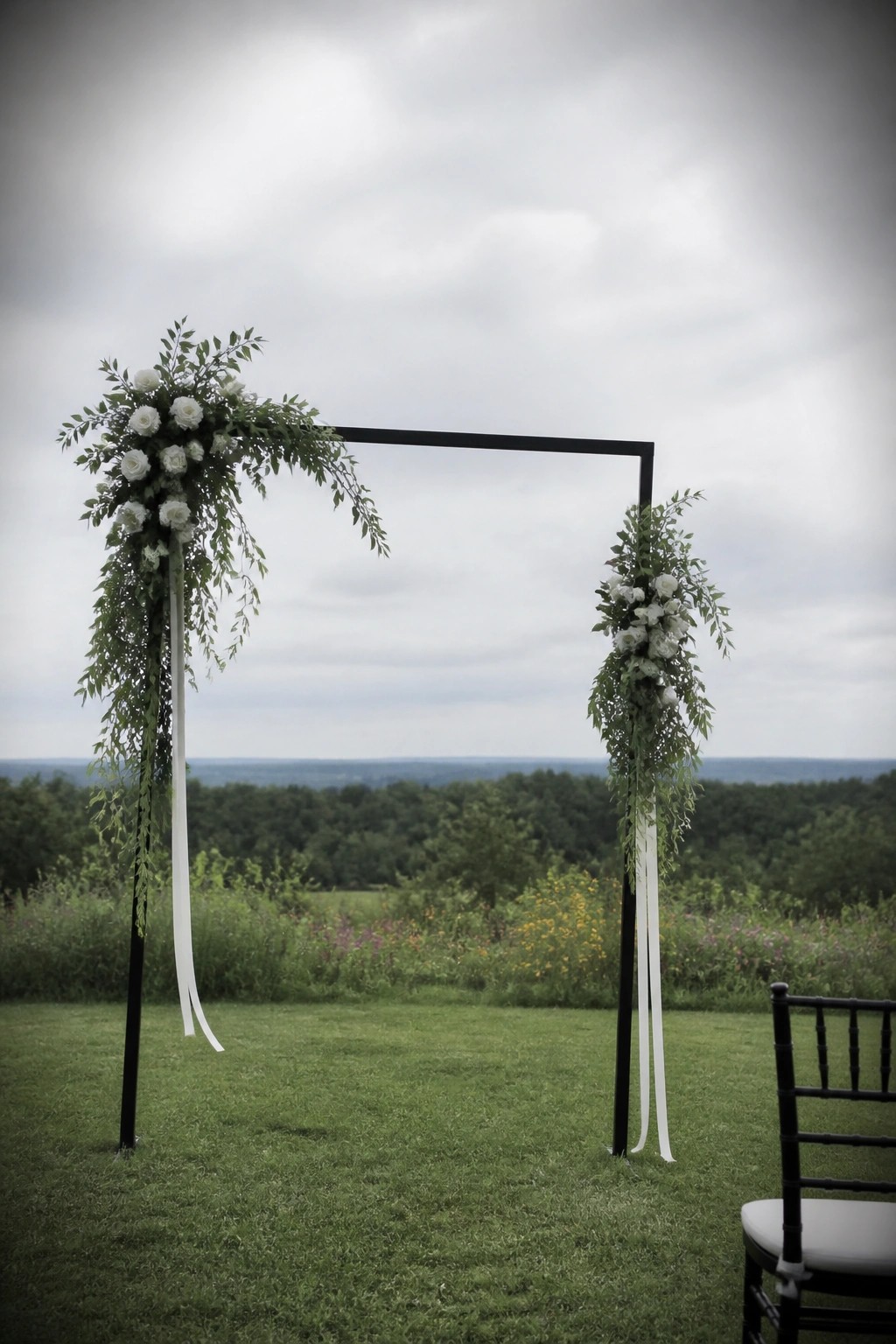 Black square wedding arch with white roses and cascading green foliage on a grassy field overlooking hills with a black chair nearby.
