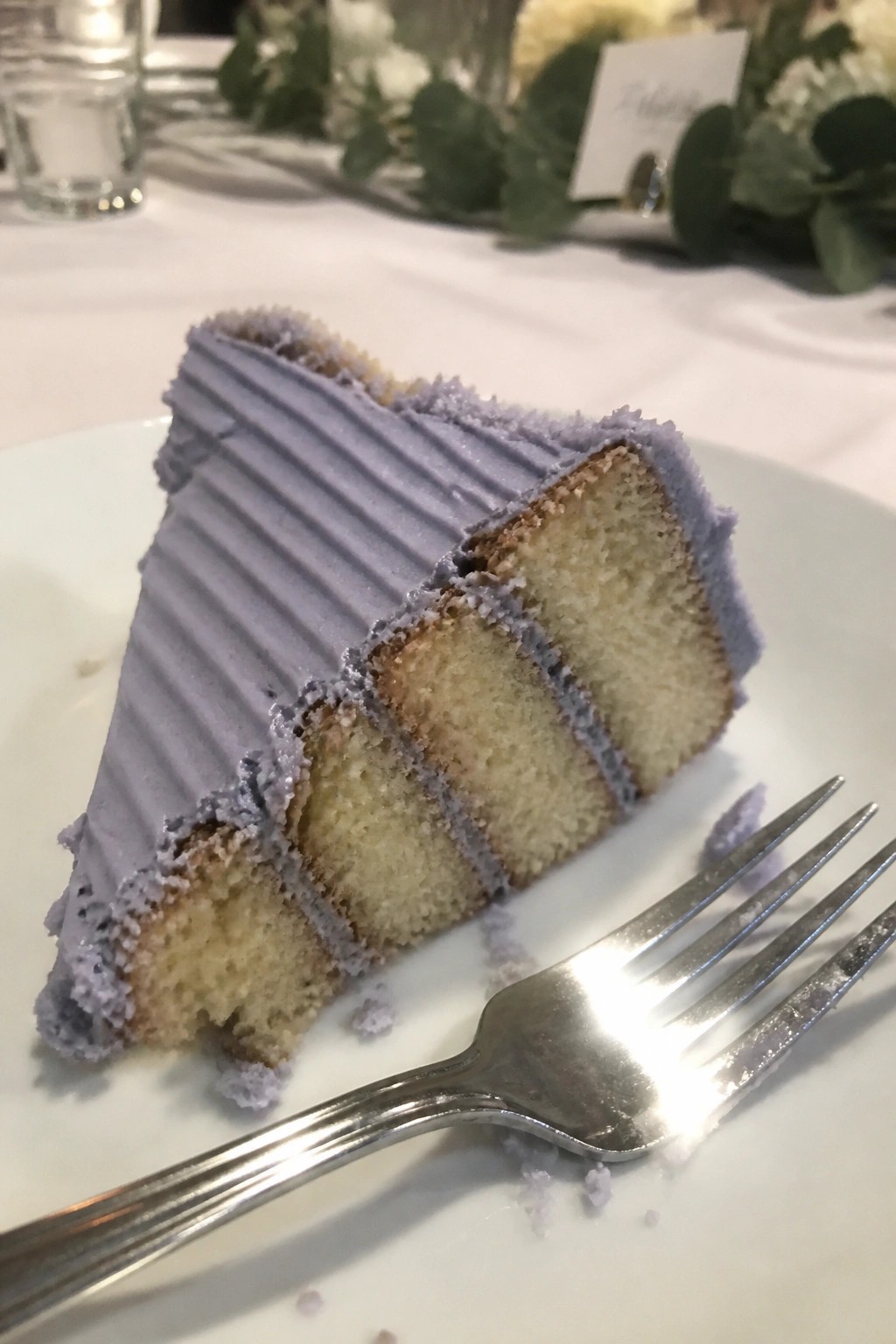 Close up of a layered vanilla cake slice with lavender purple frosting and filling on a white plate beside a fork at a wedding table