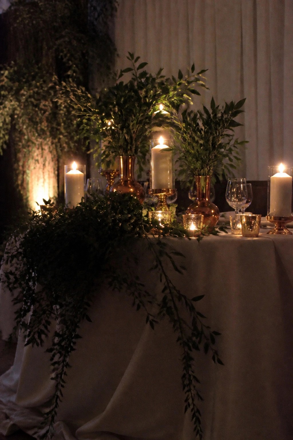 A dimly lit wedding table with white linens, cascading green foliage, copper vases holding lit white pillar candles, and scattered votive lights for a romantic evening reception.