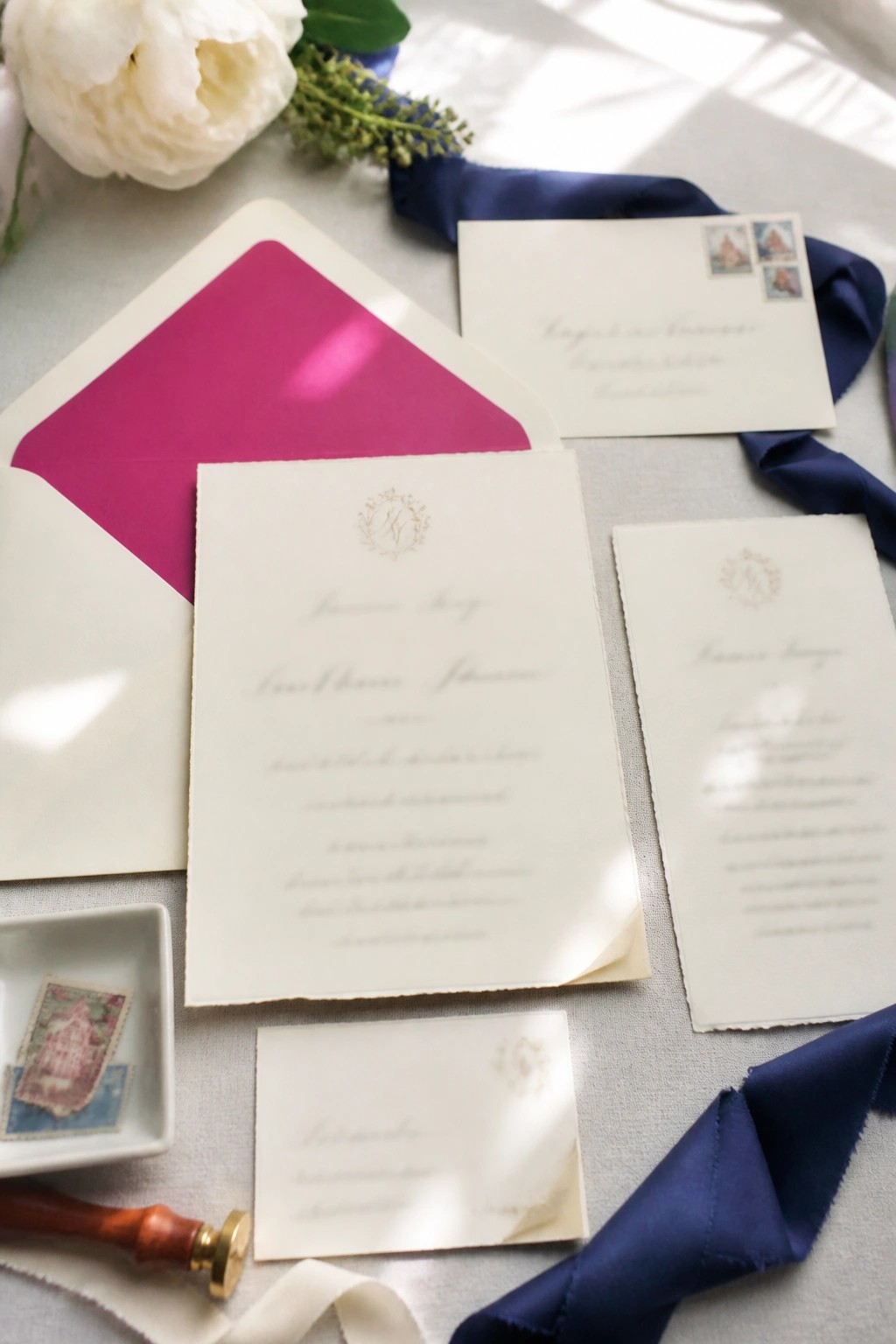 Flat lay of cream wedding invitations with magenta envelope liner, navy blue ribbon, floral monograms, vintage stamps, wax seal, and white peonies on light fabric.