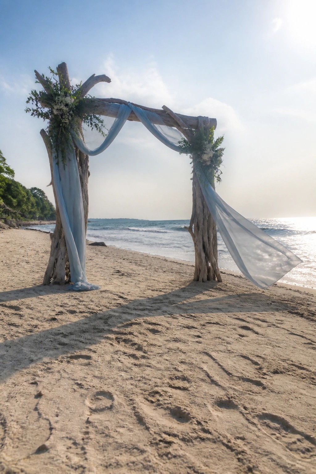Beach wedding arch made of driftwood draped in light blue fabric with white flowers overlooking the ocean on sandy shore with footprints