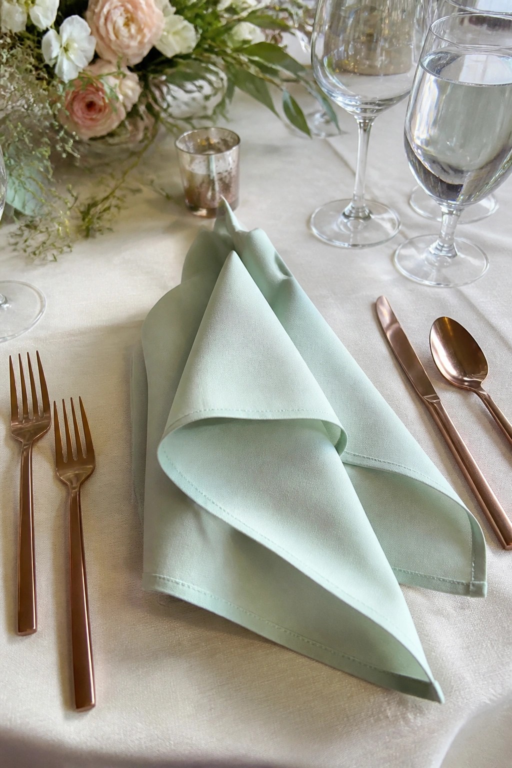 A close up of a wedding place setting with a folded mint green napkin rose gold cutlery and blush floral arrangement on a white tablecloth
