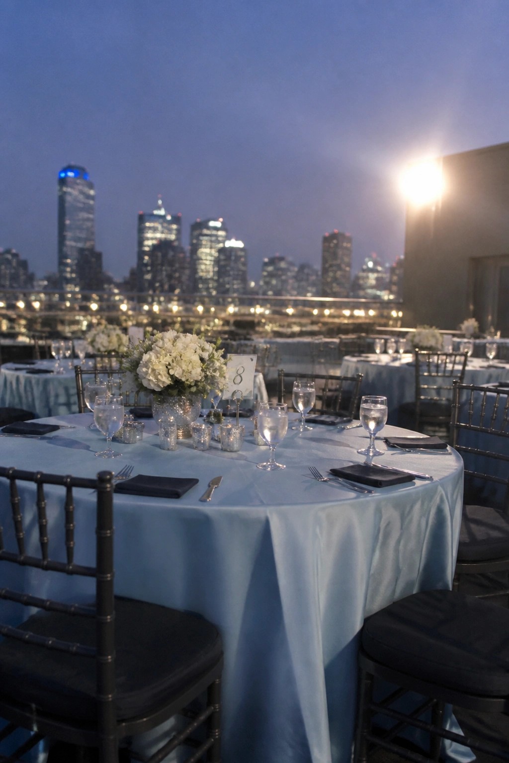 Round table with light blue tablecloth, white flowers, black napkins and chairs on a rooftop at dusk with city skyline in background