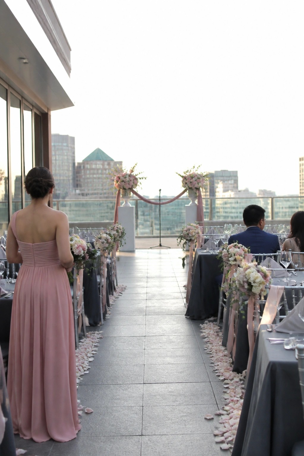 A bridesmaid in a blush pink gown stands before a rooftop wedding aisle lined with petals and gray linens topped with pink floral arrangements and a city skyline view.
