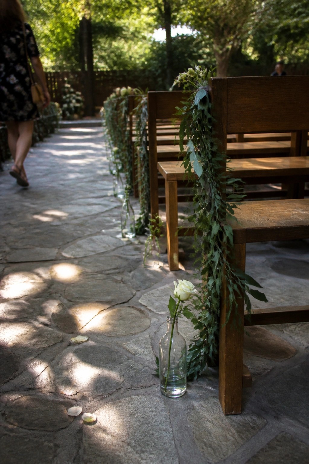 Wooden chairs with cascading green garlands line a sunlit stone aisle with rose filled bottles and scattered petals in an outdoor garden wedding
