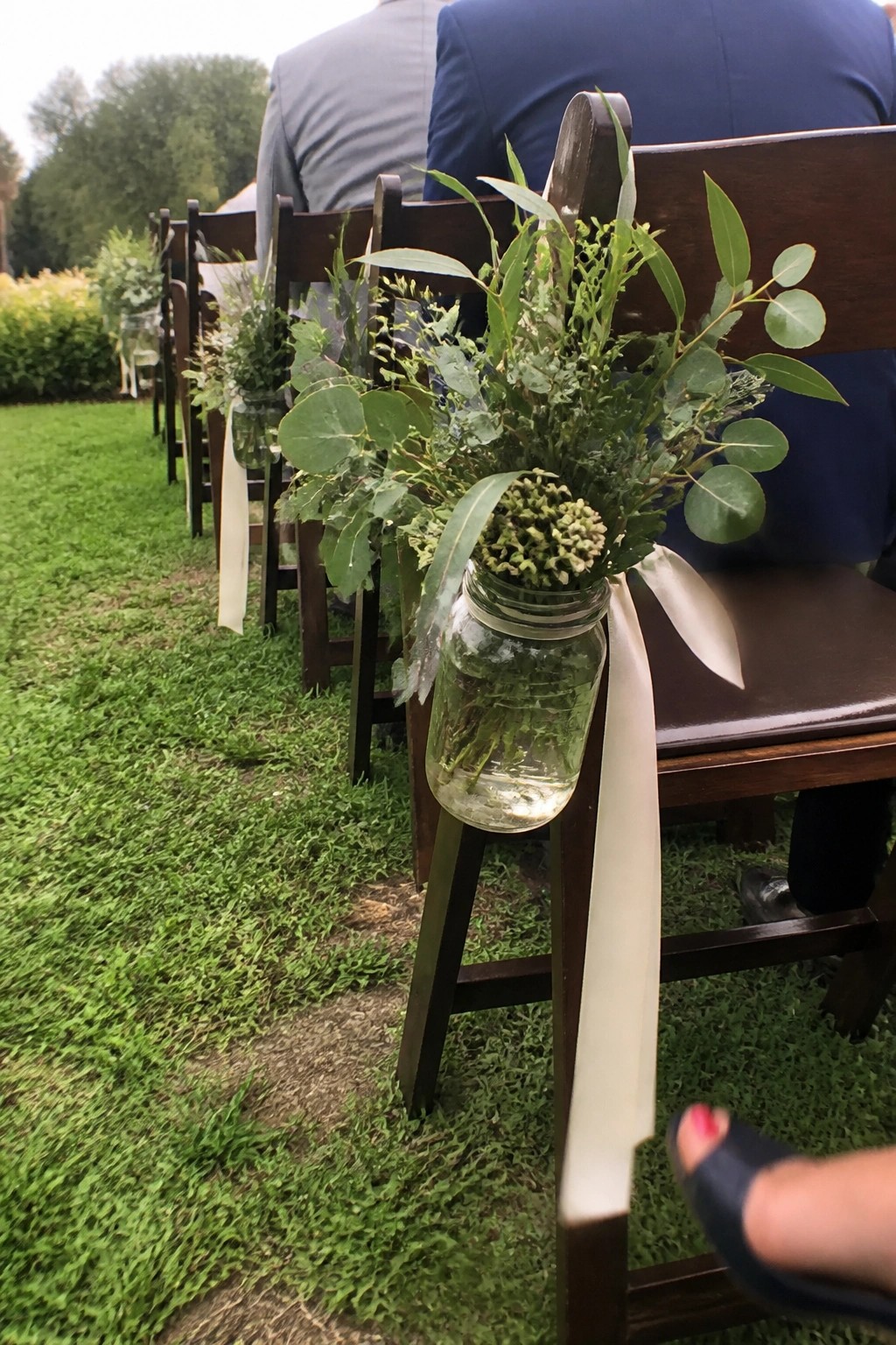 Rows of wooden chairs on grass feature mason jars with water and green foliage tied by white ribbons for an outdoor wedding ceremony.