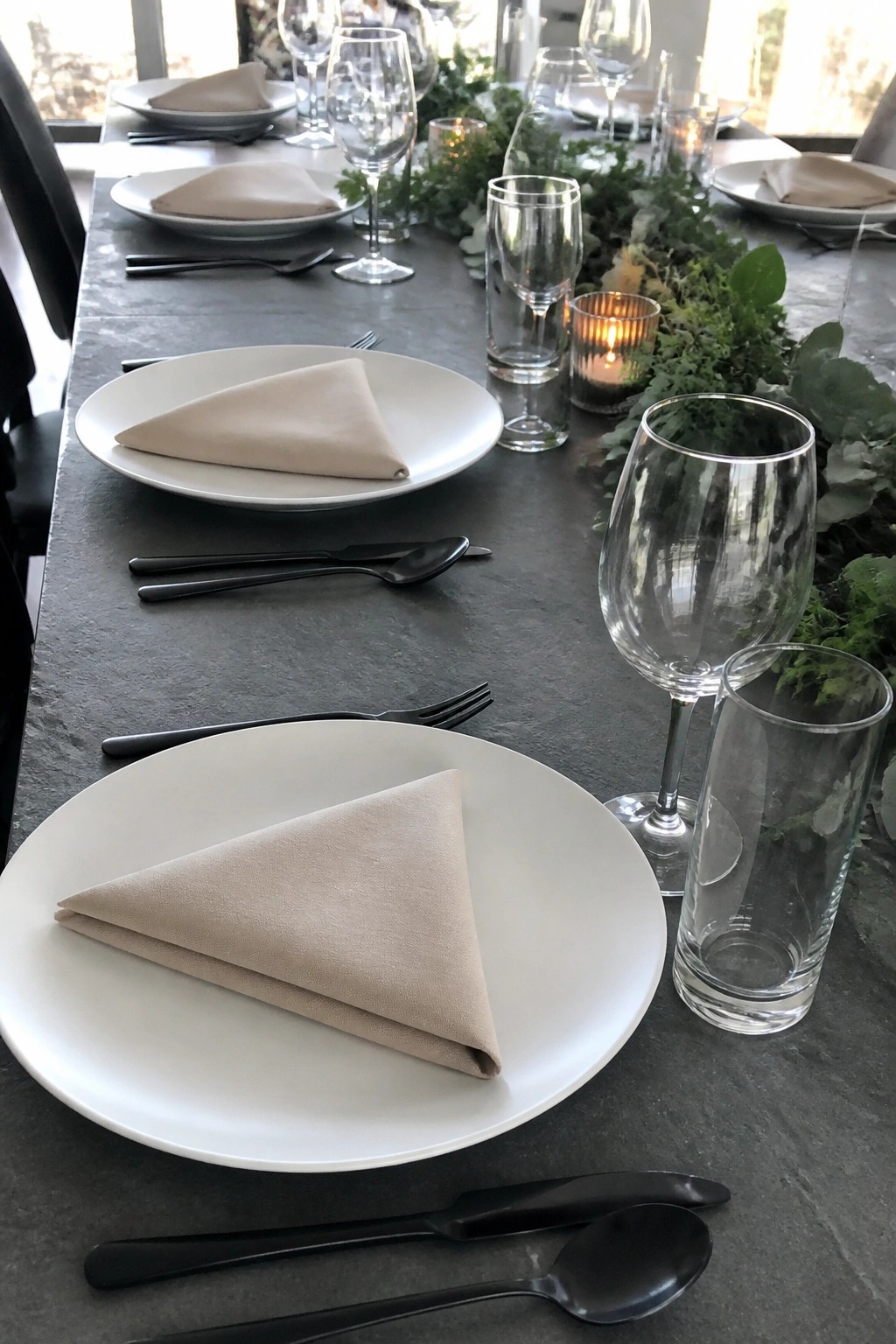 Elegant wedding reception table with white plates beige napkins black cutlery clear glassware and a green foliage centerpiece with glowing candles on a dark surface
