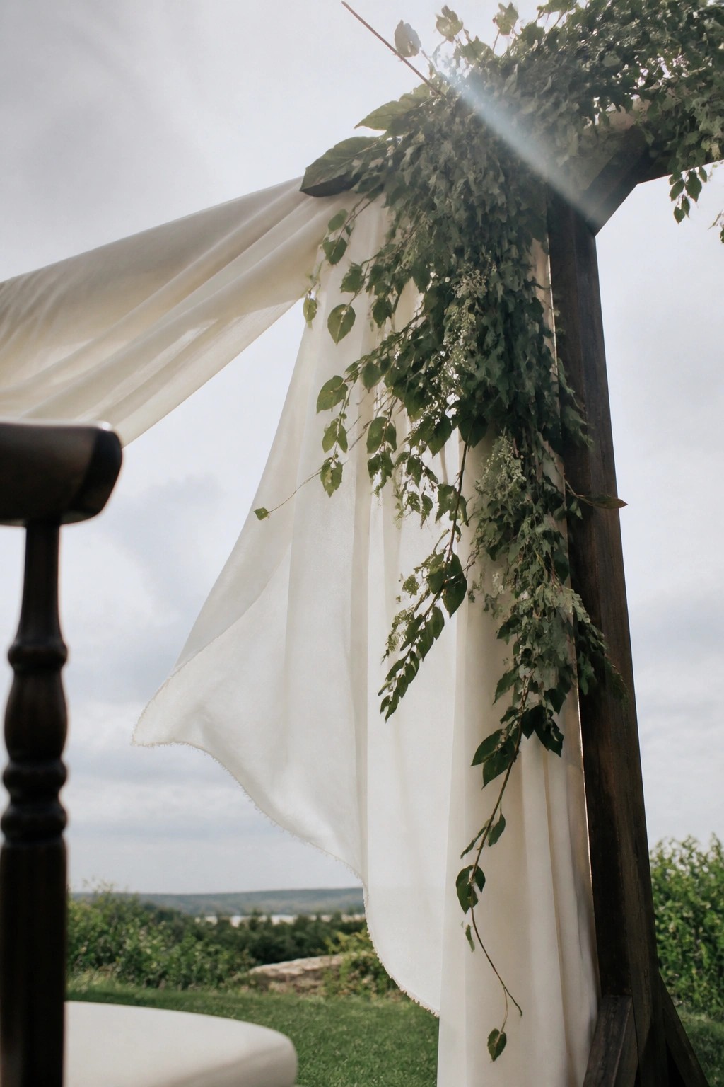 Wooden arch with cascading greenery and white fabric drapes at an outdoor wedding venue with scenic landscape background