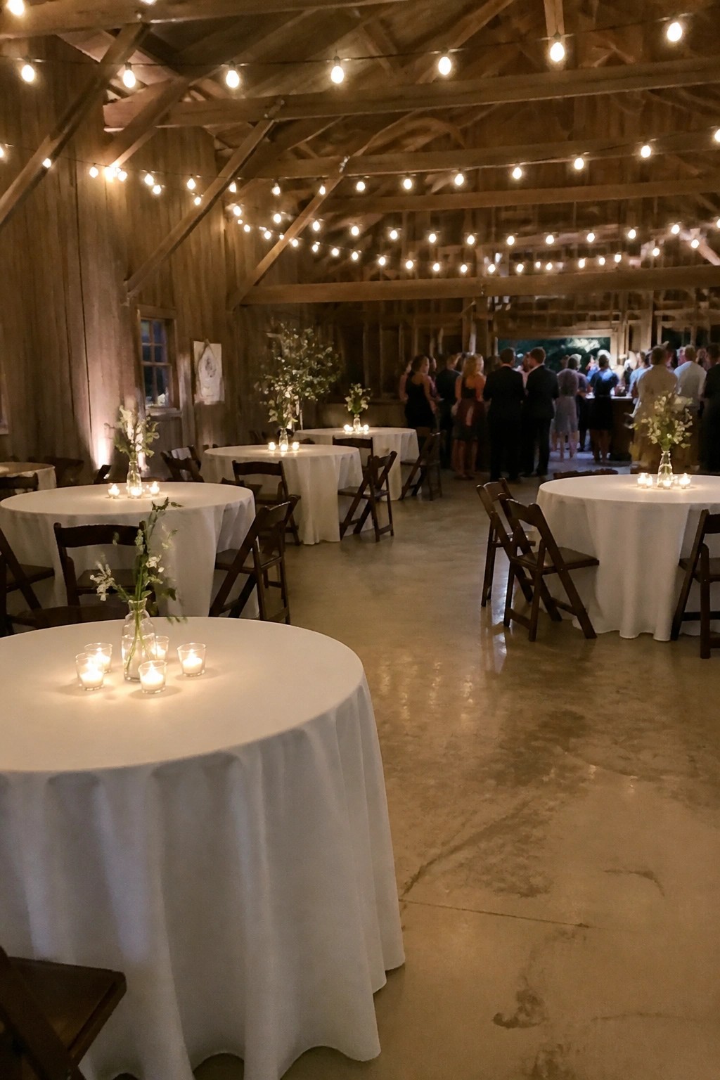 Interior of a wooden barn wedding reception with round tables covered in white linens, wooden chairs, string lights, candles, and floral centerpieces, guests mingling in the background.