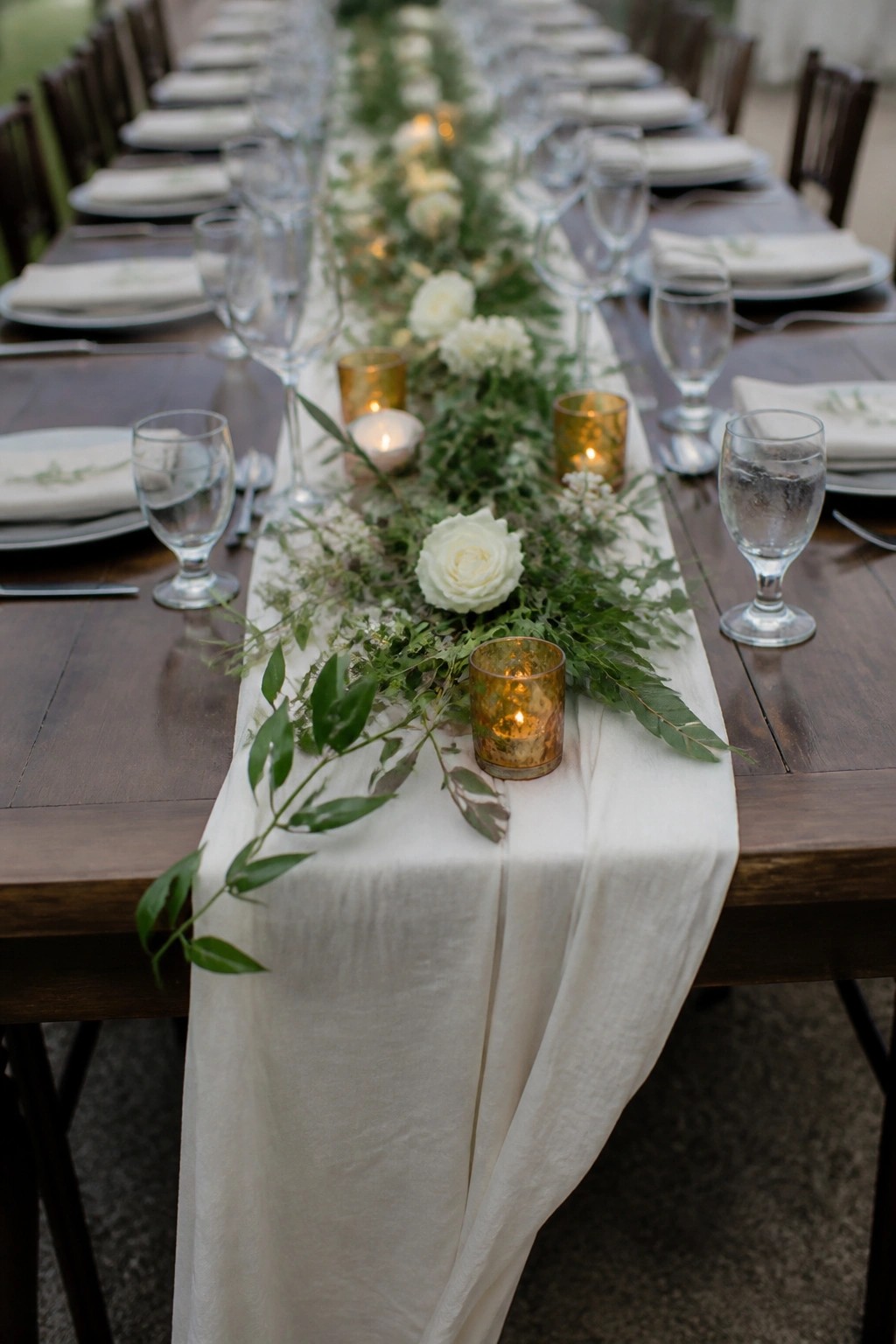 Long wooden wedding table with white runner, lush greenery, white roses, and glowing amber candle holders at an elegant outdoor reception.