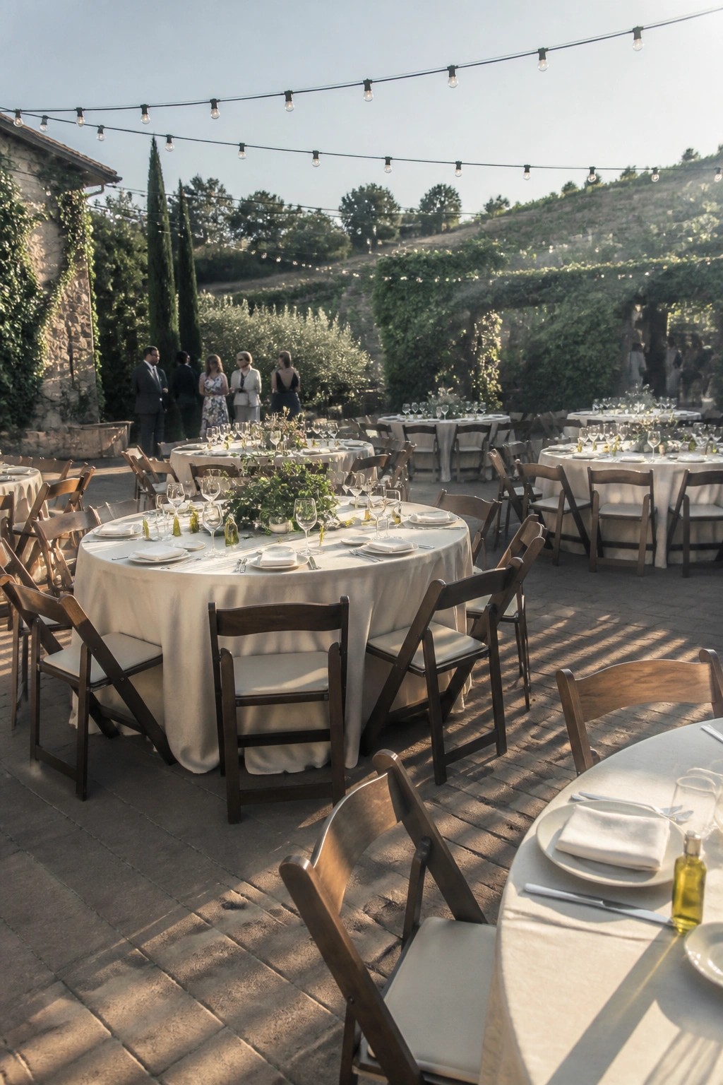 Outdoor wedding reception with round tables set in white linens and wooden chairs under string lights with vineyard hills in the background
