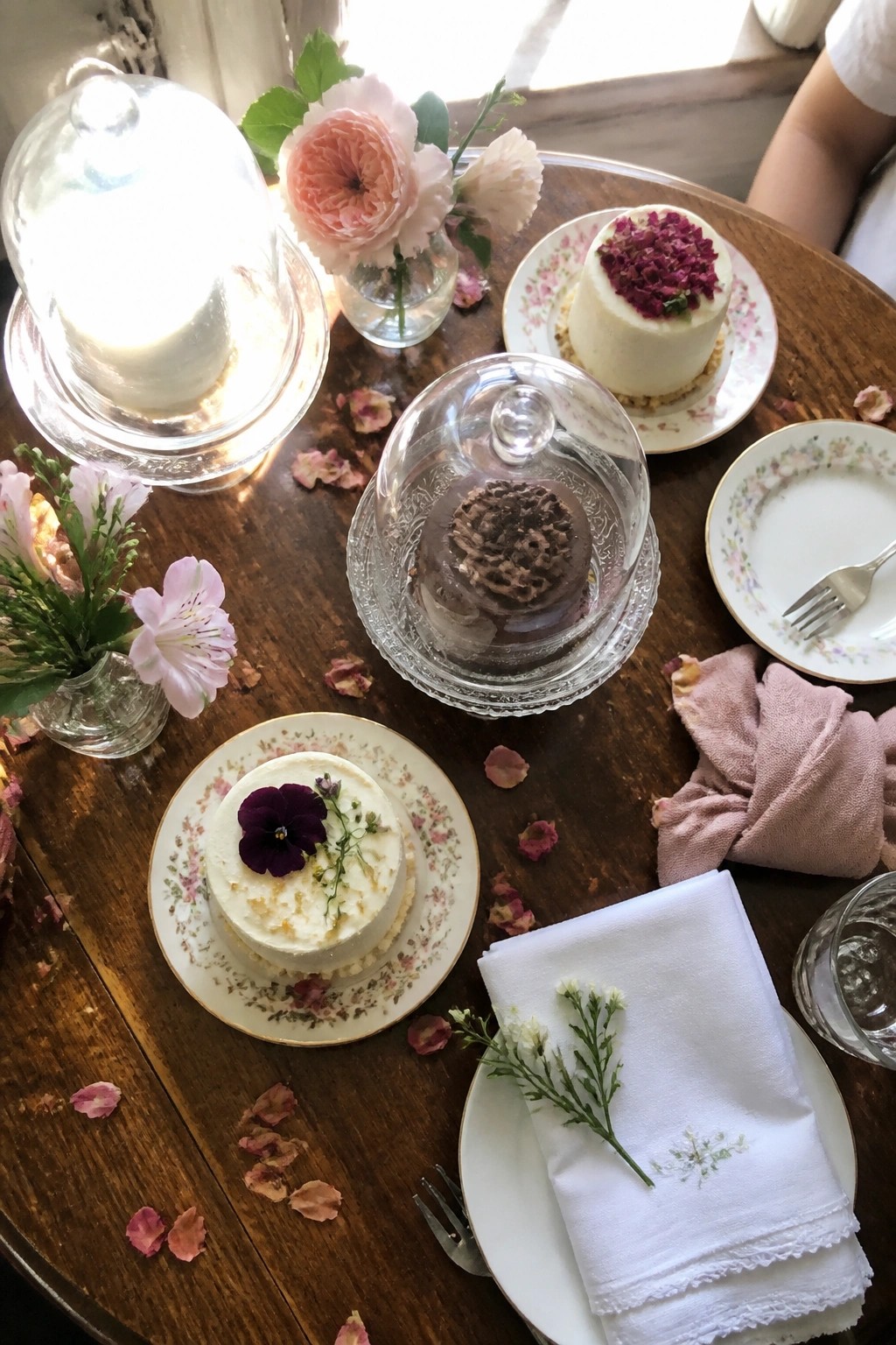 Top down view of a wooden table with three miniature cakes under glass cloches, pink flowers, a lit candle, rose petals, and elegant plates in a romantic intimate wedding setting.