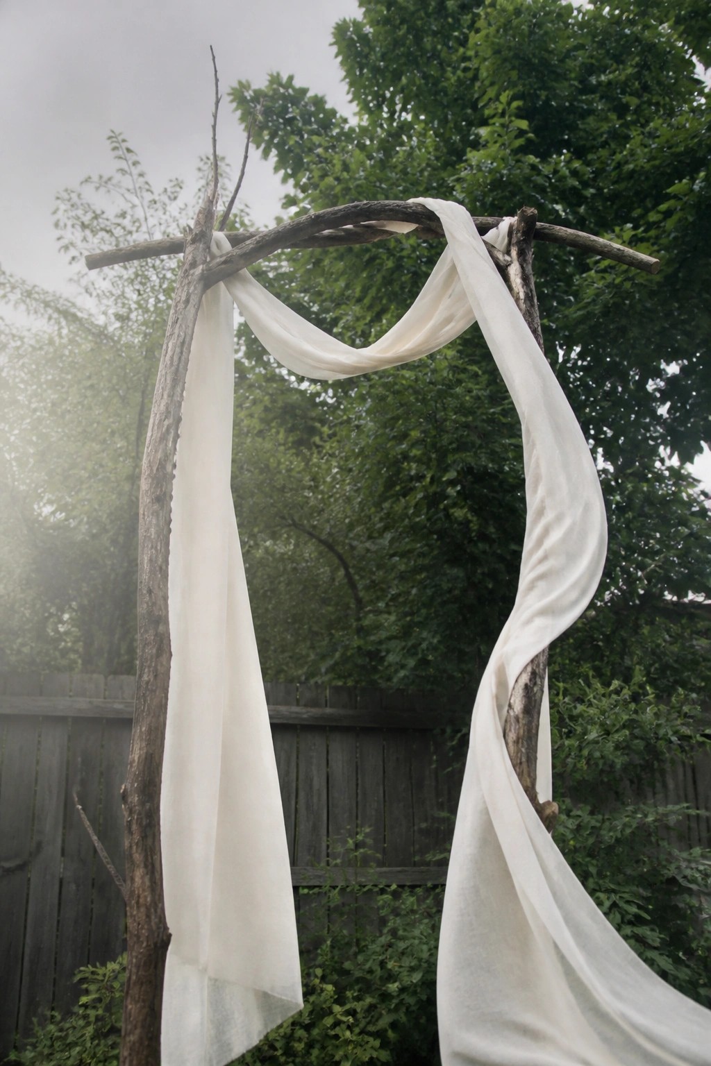 A rustic wedding arch made of tree branches draped with flowing white fabric stands in a garden with green trees and a wooden fence in the background.