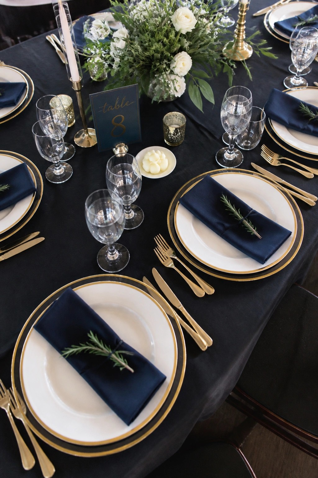 Top down view of an elegant black tie wedding table with black linens, white and gold plates, navy napkins, gold cutlery, and white rose floral centerpieces.