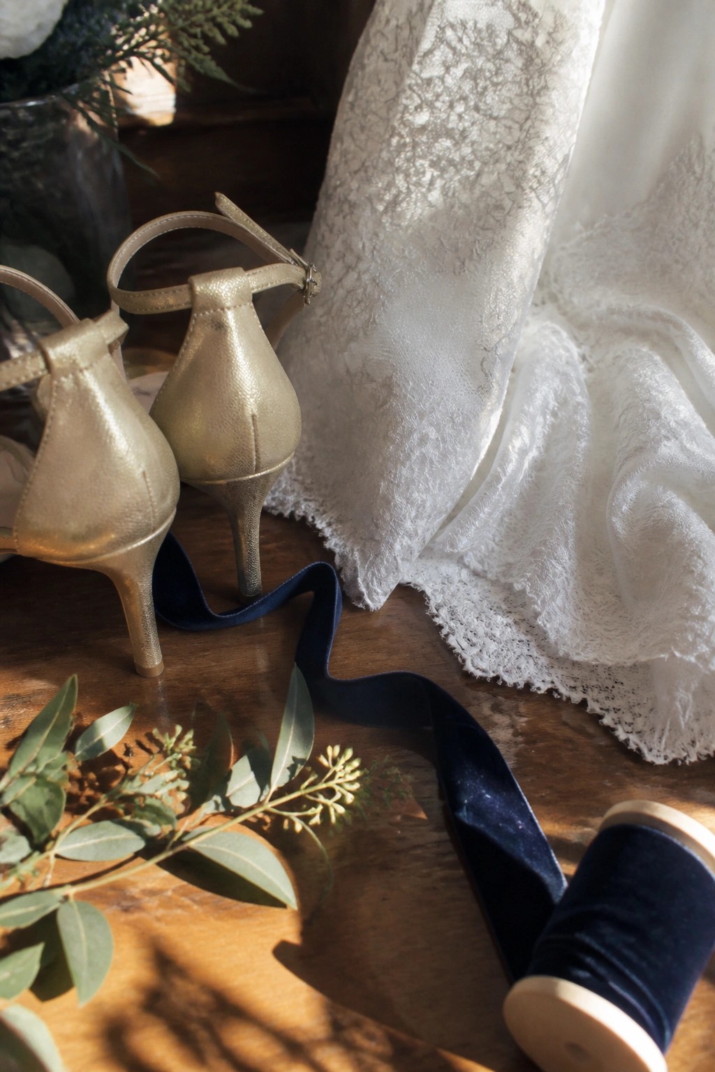 Close up of gold high heeled wedding shoes next to white lace fabric, a navy velvet ribbon spool, and green leaves on a wooden table in soft light.