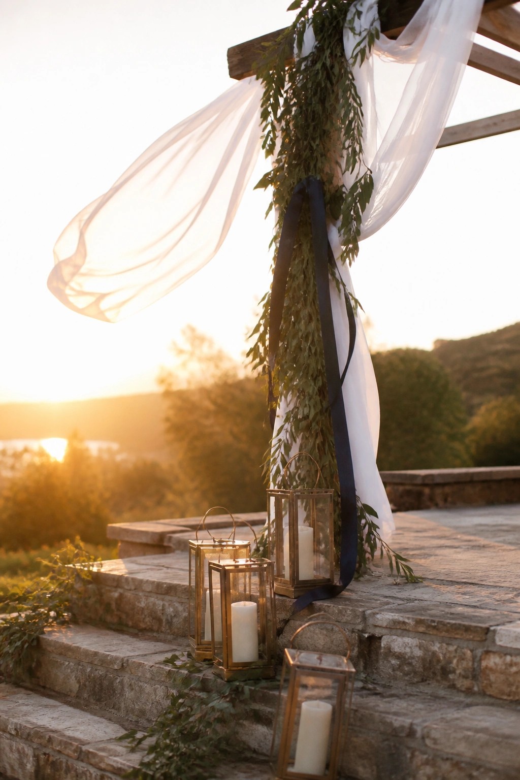 Stone steps at sunset lined with gold lanterns holding white candles and accented by navy ribbon and green foliage on a wooden arch draped in white fabric.