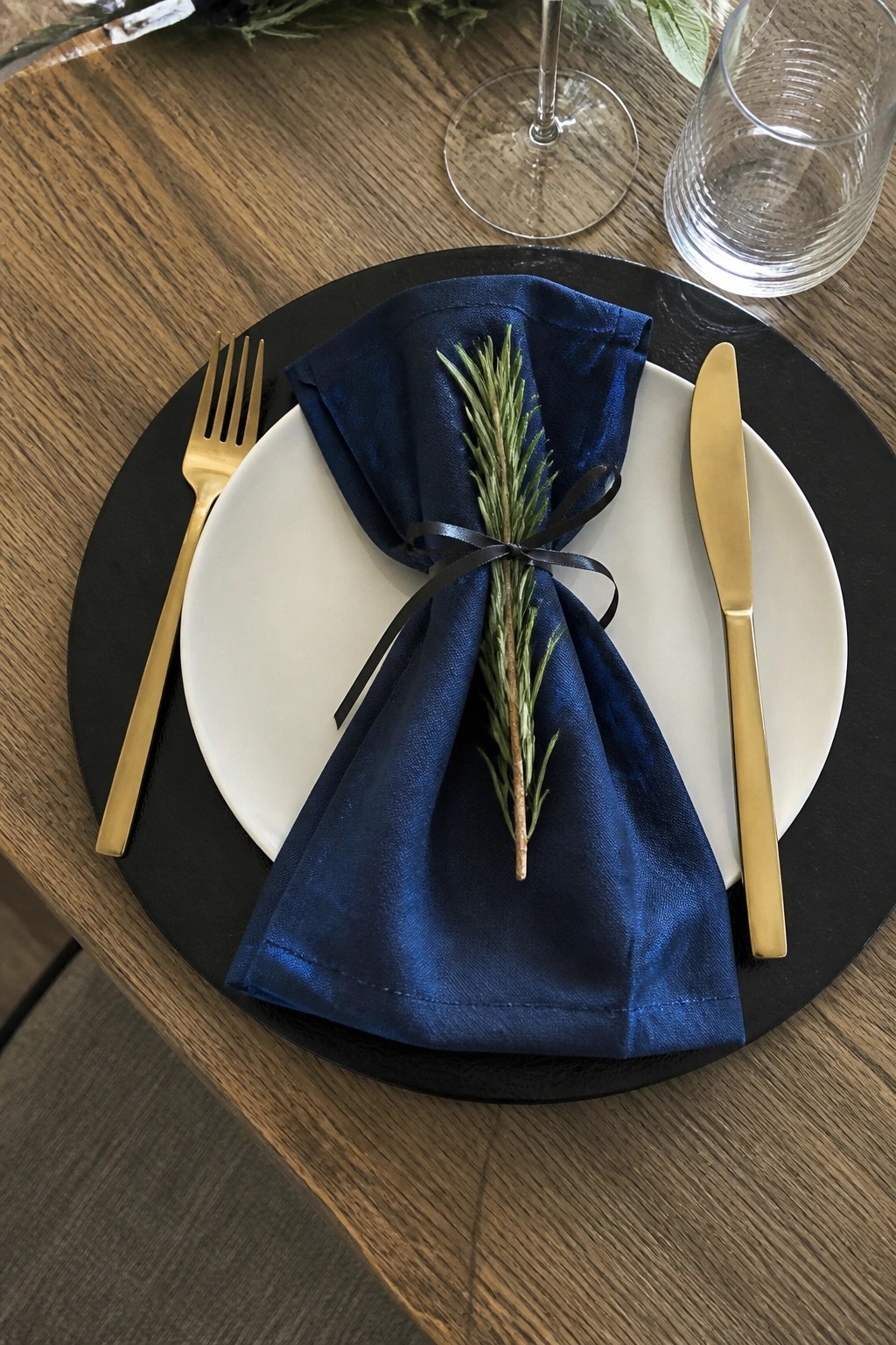 Close up of a black charger plate with white dinner plate, navy blue folded napkin tied with black ribbon holding rosemary, gold fork and knife, and clear glasses on a wooden table.