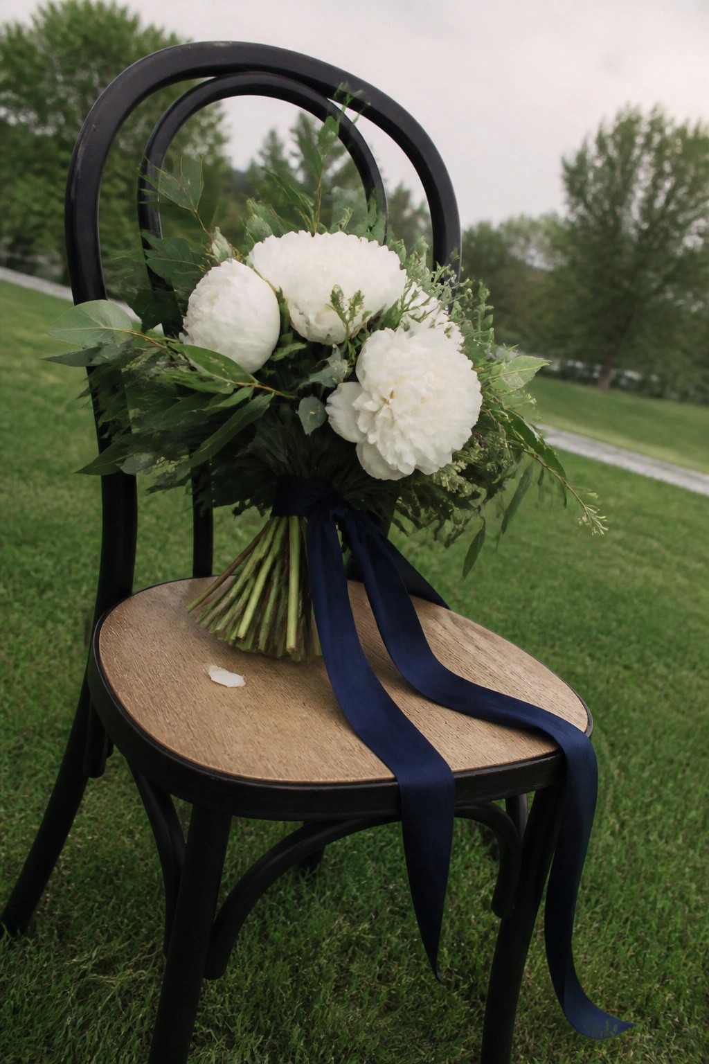 A bouquet of white peonies and greenery tied with a navy ribbon sits on a black chair outdoors.