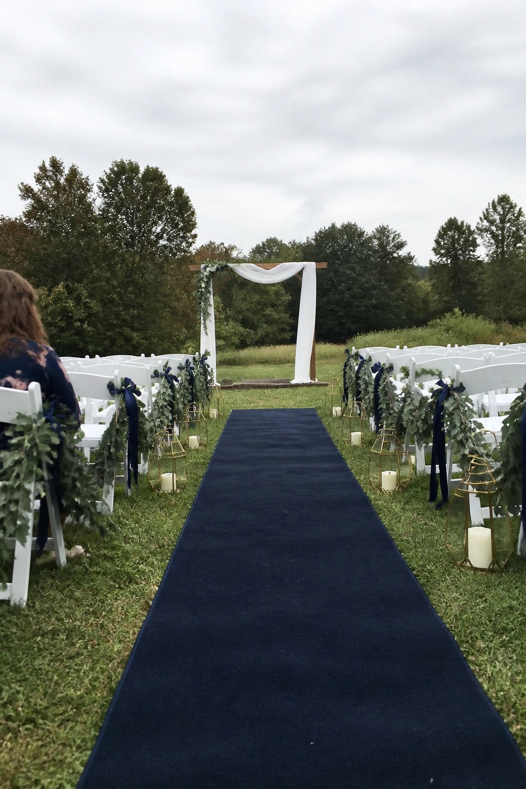 Outdoor wedding aisle with navy runner, white chairs with navy and green decor, gold candle lanterns, and a white draped arch under overcast skies.